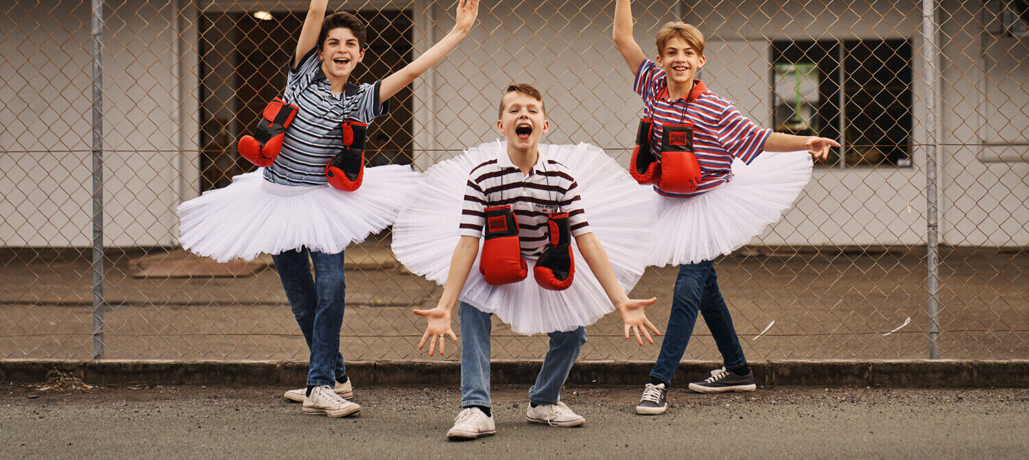 Three young actors playing Billy Elliot on the Gold Coast do a ballet pose wearing a tutu over their regular clothes with boxing gloves around their necks